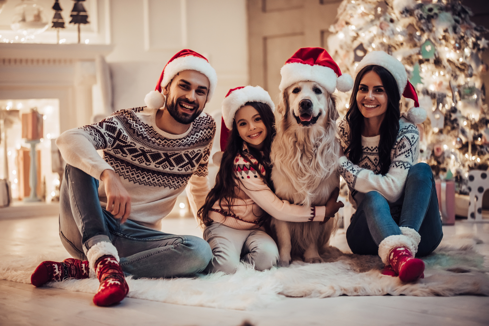 A family of three, wearing Santa hats and festive sweaters, sits on a cozy rug beside a decorated Christmas tree. They are smiling and posing with their golden retriever, who also wears a Santa hat, celebrating another healthy year thanks to their trusted veterinarian.