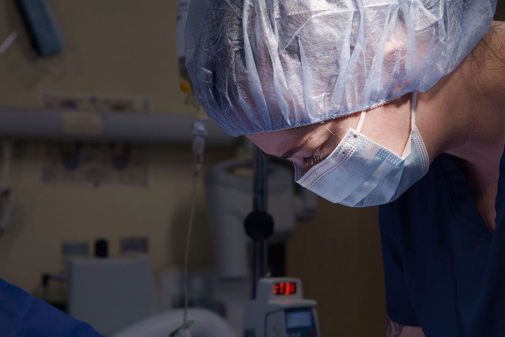 A dedicated healthcare professional in scrubs, surgical cap, and mask leans over with the precision of a vet focused on a procedure. Medical equipment is visible in the background, highlighting a clinical or hospital setting.