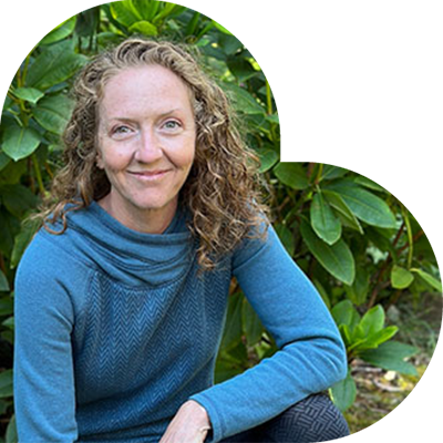 A person with curly hair, who happens to be a dedicated veterinarian, is wearing a blue sweater and posing outdoors in front of lush green foliage. They are smiling and sitting in a relaxed manner.